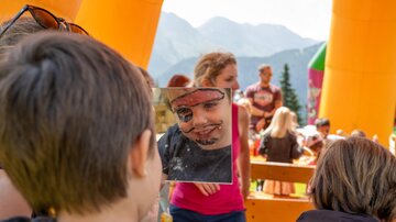 A child looks into a mirror and sees a face with makeup. In the background, other people and a colorful atmosphere in the mountains can be seen.