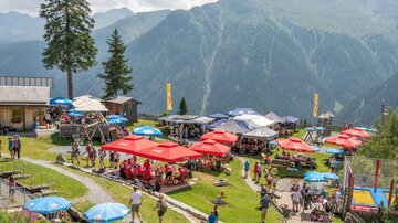 A lively mountain lodge with terraces under red and blue sun umbrellas. In the background, impressive green mountains and a clear sky can be seen.