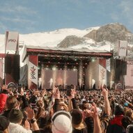 Ein großes Musikfestival in den Bergen mit vielen Menschen, die zur Show tanzen. Im Hintergrund sind schneebedeckte Berge und eine Bühne mit Lichtern zu sehen.