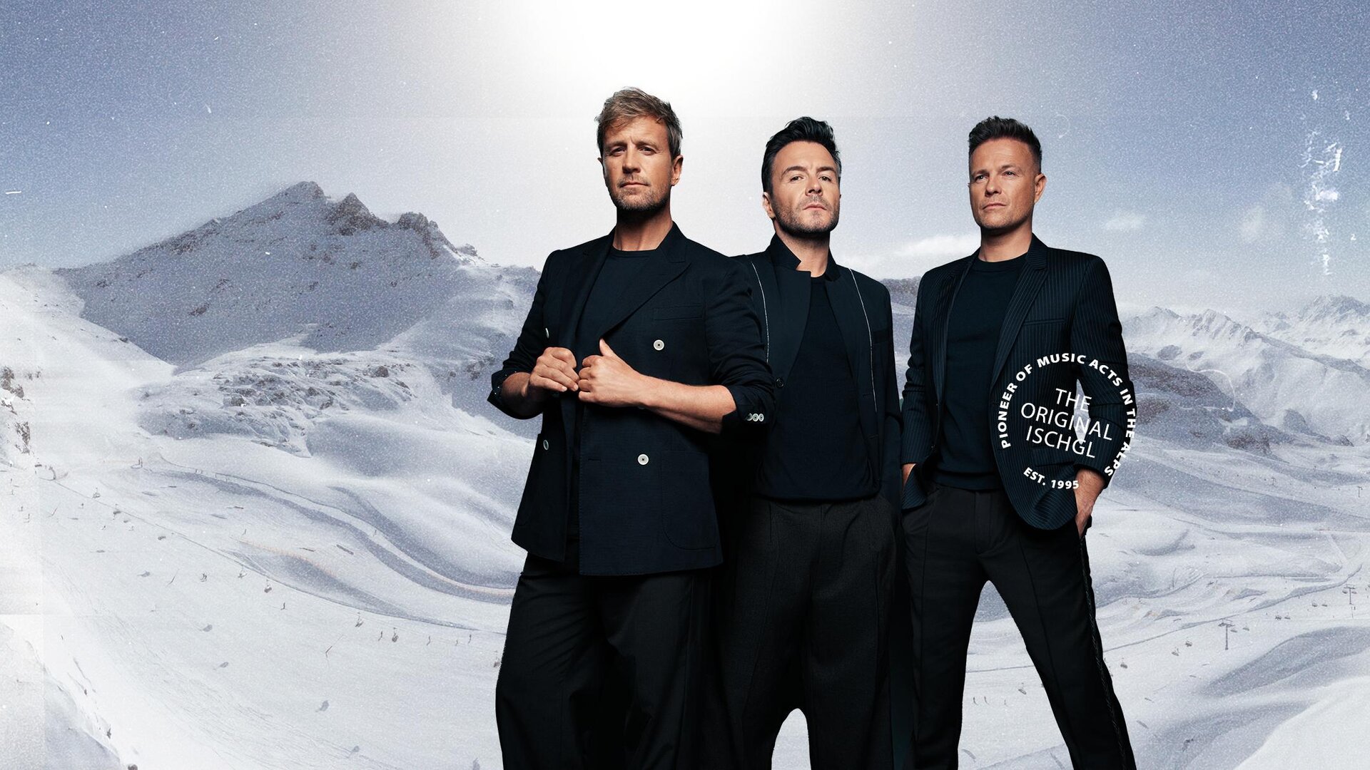 Three men stand in front of a snowy mountain landscape. They are wearing elegant black clothing and looking at the camera.