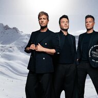 Drei Männer stehen vor einer verschneiten Berglandschaft. Sie tragen elegante schwarze Kleidung und blicken in die Kamera.