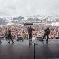Eine große Menschenmenge genießt ein Konzert in den Bergen. Im Hintergrund sind schneebedeckte Gipfel und ein Veranstaltungsgebäude zu sehen.