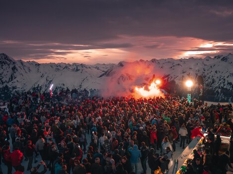 Eine große Menschenmenge versammelt sich in den Bergen bei Sonnenuntergang. Feierliche Stimmung mit bunten Lichtern und Rauchfackeln.
