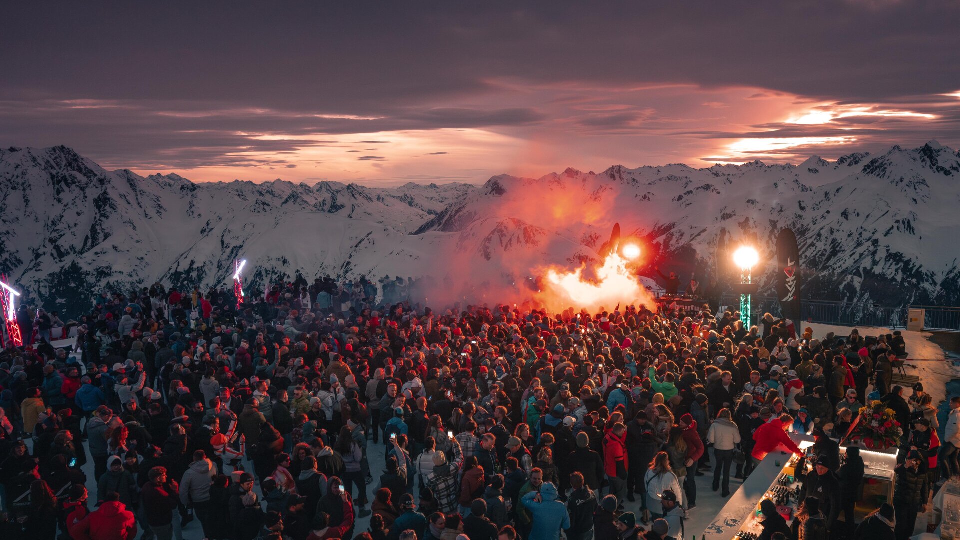 Eine große Menschenmenge versammelt sich in den Bergen bei Sonnenuntergang. Feierliche Stimmung mit bunten Lichtern und Rauchfackeln.