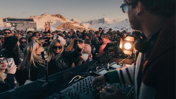 Eine lebhafte Party im Freien mit vielen Menschen und einem DJ. Im Hintergrund sind schneebedeckte Berge zu sehen.