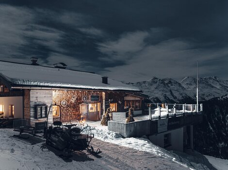 Eine Holzski-Hütte im Schnee mit einem atemberaubenden Bergpanorama. Die Szenerie ist bei Nacht mit einem klaren Himmel und einem beleuchteten Innenbereich.