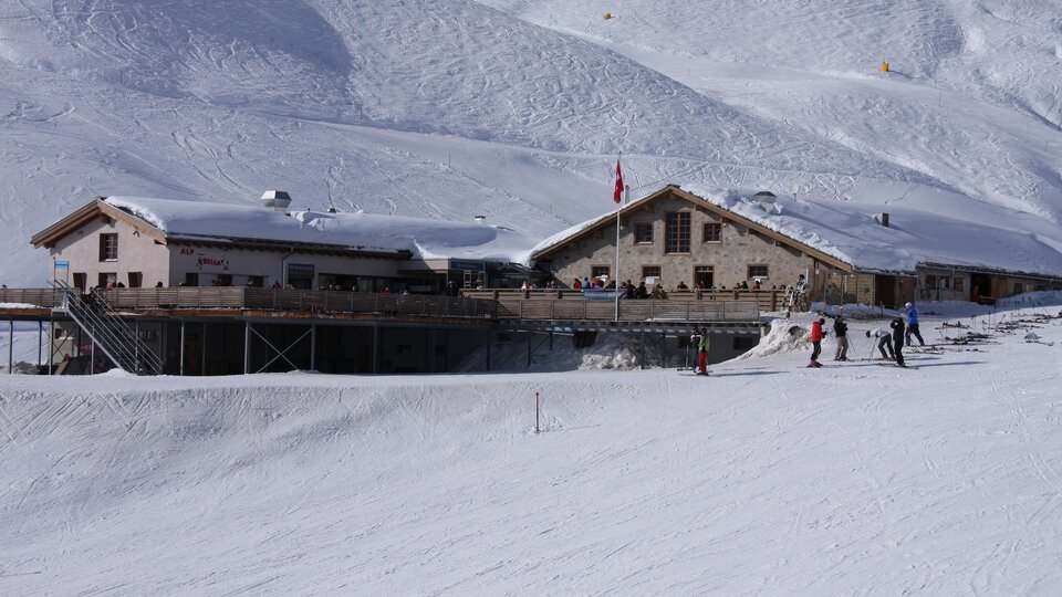 A mountain hut in the snow with skiing people. The landscape is surrounded by snow-covered mountains.