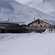 Eine Berghütte im Schnee mit skifahrenden Personen. Die Landschaft ist von schneebedeckten Bergen umgeben.
