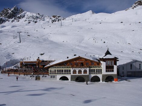 A snow-covered landscape with mountain ranges in the background. In the foreground stands a traditional mountain house.