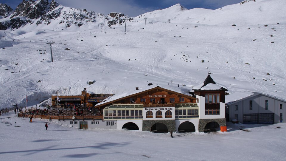 A snow-covered landscape with mountain ranges in the background. In the foreground stands a traditional mountain house.