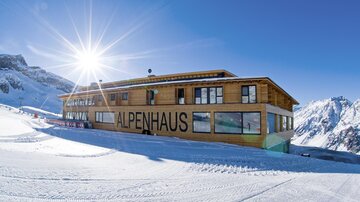 A modern wooden building with the inscription "ALPENHAUS" in a snowy mountain landscape. The sun is shining brightly in the clear blue sky.
