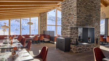 A modern restaurant with large windows and a view of snow-covered mountains. The decor consists of wooden elements and cozy red chairs.
