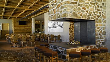A modern restaurant with wooden decor and a distinctive stone wall. In the center, there is an open fireplace, surrounded by cozy seating.