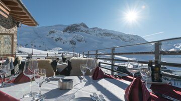 A cozy restaurant terrace with a view of snow-covered mountains and bright sunshine. The table is festively set with glasses and napkins.