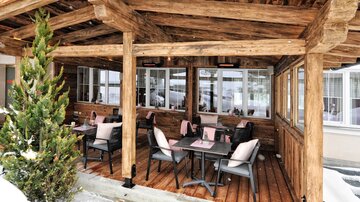 A cozy terrace with wooden beams and tables. The surroundings are wintry, with snow and a green plant.