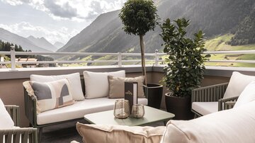 A cozy terrace with modern seating and plants. In the background, majestic mountains and a clear sky can be seen.