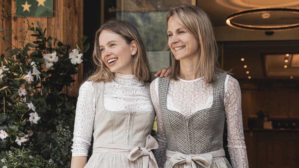 Two women are smiling in front of a building. Both are wearing traditional dresses with modern accents.