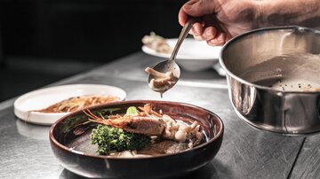 A chef serves a dish in an elegant bowl. The presentation includes shrimp, vegetables, and a sauce.