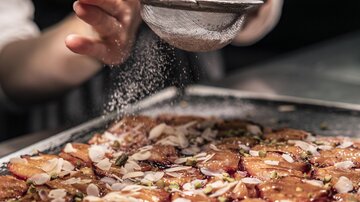 A hand sifts sugar over a tray of sweet, topped plates. The top is adorned with almonds and other ingredients.
