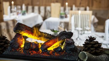 A cozy dining area with table decorations and candles. In the foreground, decorative fireplace logs are burning.