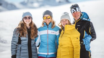 A group of four people stands in a snowy mountain area. They are all wearing warm winter jackets and hats, and the sun is shining brightly.