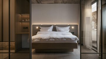 A modern bedroom with a large double bed and soft lighting. The walls are in neutral tones, and there are large windows that let in natural light.