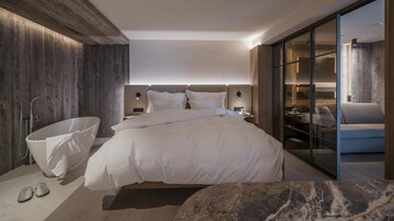 A modern bedroom with a large bed and elegant wooden design. A freestanding bathing area and large windows provide pleasant light.