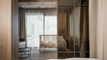 A modern room with wooden paneling and large windows. In the background, there is a baby crib, and the room feels bright and inviting.