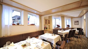 An elegant restaurant interior with white tablecloths and set tables. Large windows let in plenty of light and provide a view of the outdoors.