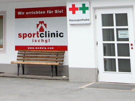 A sports clinic with a red sign and a bench outside. Next door is a pharmacy sign.