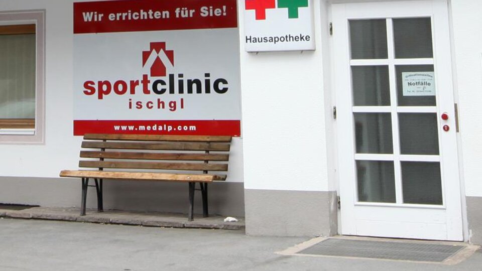 A sports clinic with a red sign and a bench outside. Next door is a pharmacy sign.