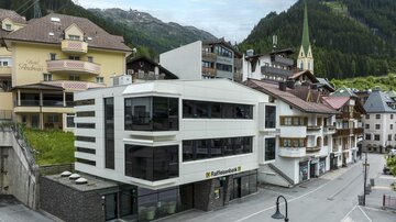Ein modernes Gebäude mit großen Fenstern in einer malerischen Landschaft. Im Hintergrund sind traditionelle Häuser und ein Kirchturm zu sehen.