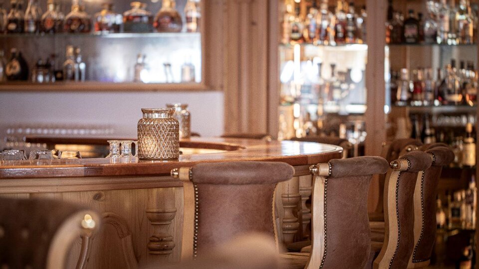Eine stilvolle Bar mit holzverkleideten Stühlen und eleganten Gläsern. Im Hintergrund sind Regale mit verschiedenen Spirituosen sichtbar.