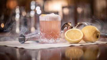 Ein eleganter Cocktail in einem Glaskelch, umgeben von dampfendem Eis. Neben dem Getränk liegen frische Zitronenhälften auf einem weißen Tisch.