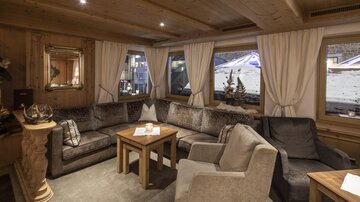 Ein gemütlicher Wohnbereich mit einem großen, U-förmigen Sofa und einem Holztisch. Durch die Fenster sieht man eine winterliche Landschaft.