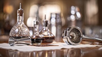 Eine elegante Bar mit verschiedenen Gläsern und Utensilien für die Cocktailzubereitung. Im Vordergrund sind ein Milchschäumer und ein Messbecher zu sehen.