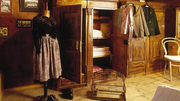A cozy room with an old wardrobe, a doll in traditional attire, and various pieces of clothing hanging from it. The wooden floor and walls give the room a warm atmosphere.