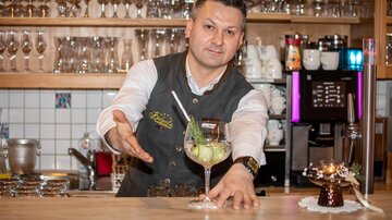 A bartender proudly presents a colorful cocktail at the bar. In the background, numerous glasses and a well-stocked bar are visible.