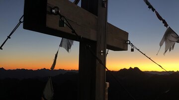 A wooden cross at the mountain peak, silhouetted against the sunset. Colorful flags flutter in the wind.