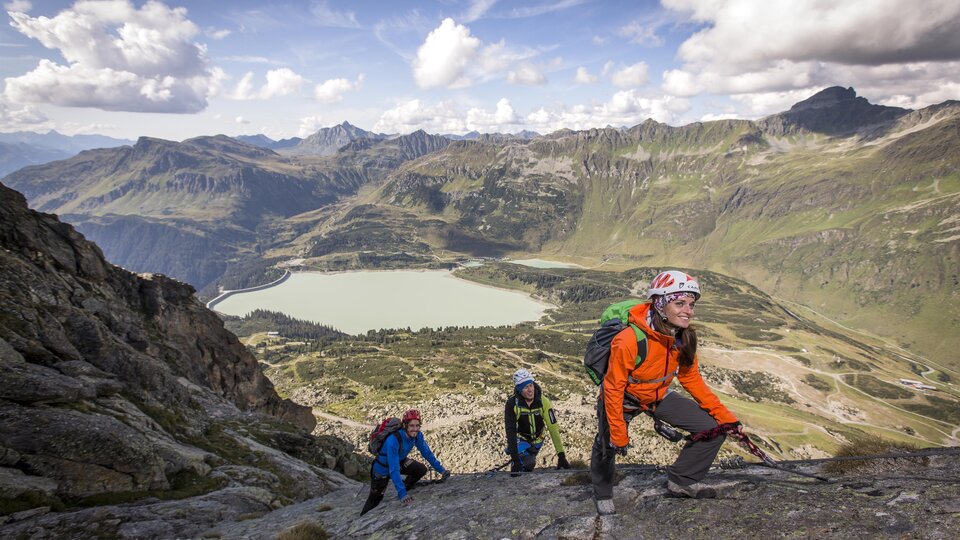 A group of climbers is ascending a rock in the Alps. In the background, a clear lake and impressive mountain landscapes can be seen.