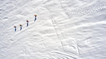 Four people are hiking across an untouched snowy landscape. Their footprints are clearly visible in the white snow.
