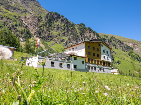 A cozy building on a meadow, surrounded by tall mountains. The sky is clear and blue, creating a calm atmosphere.