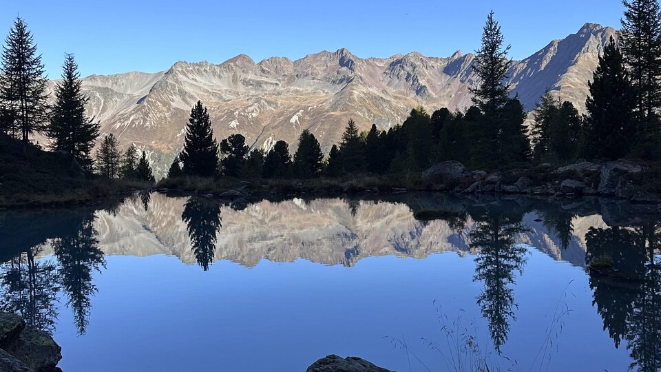 Ein ruhiger Bergsee umgeben von Tannen und Bergen. Die Reflektionen sorgen für eine malerische Aussicht unter einem klaren blauen Himmel.