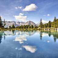 Ein ruhiger Bergsee mit kristallklarem Wasser und spiegelnden Wolken. Die umliegenden Bäume und Berge schaffen eine idyllische Naturszene.