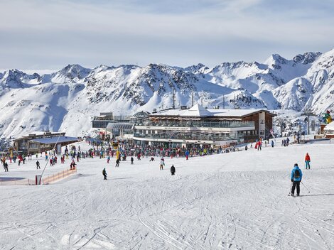 A lively ski resort with many people on the slopes. In the background, there are snow-covered mountains and a modern building.
