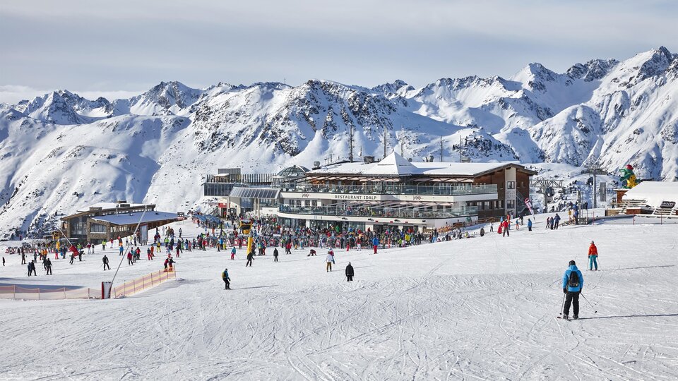 A lively ski resort with many people on the slopes. In the background, there are snow-covered mountains and a modern building.