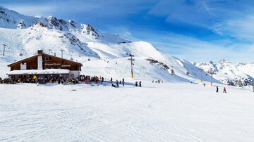 A snow-covered mountain with a ski hut and many people. In the background, majestic mountains are visible under a clear sky.