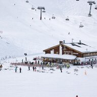 Ein Holzhaus in den Bergen, umgeben von Schnee und Skifahrern. Im Hintergrund sind Skilifte zu sehen.