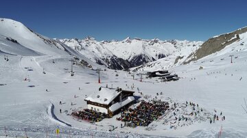 A snow-covered mountain landscape with a ski lodge in the foreground. Many skiers are on the slopes, and the peaks are clear and sunny.
