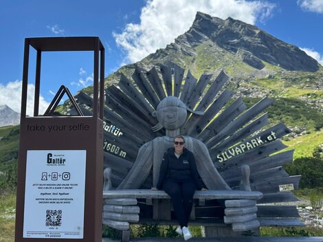 A person is sitting on a large wooden figure wearing sunglasses. In the background, there are mountains and a clear blue sky.